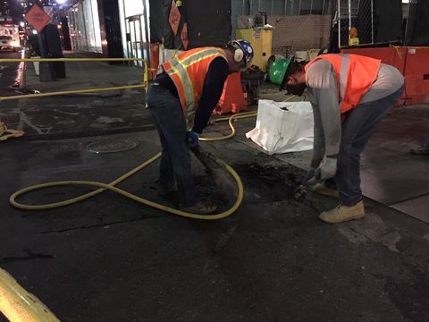 Construction workers in safety vests hand-digging a street trench at a utility work site in New York City.